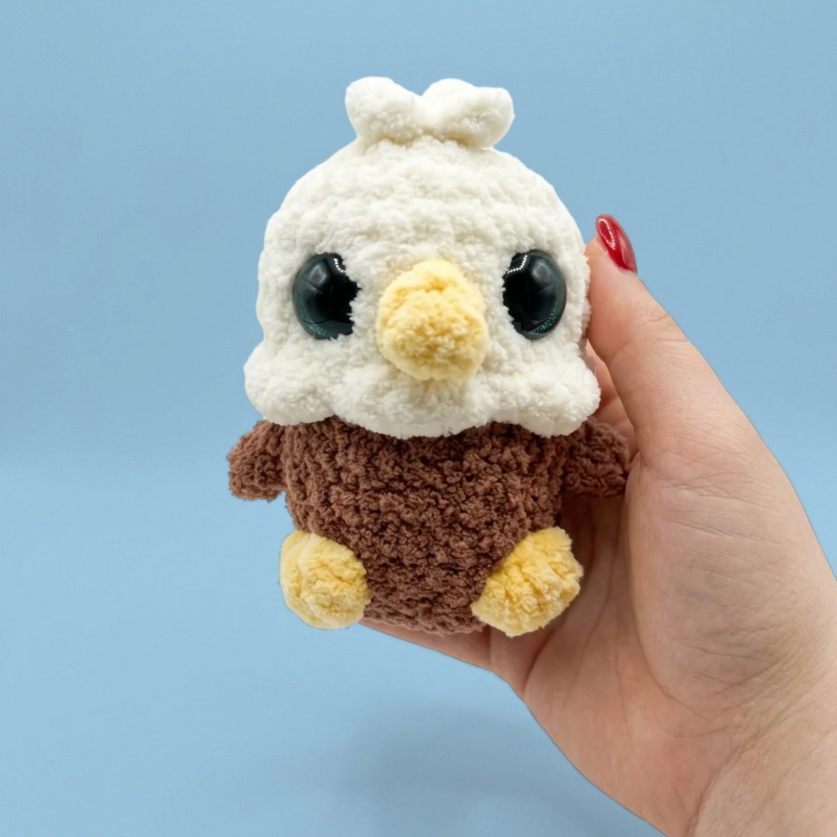A small crocheted plush toy shaped like a bald eagle held in a person's hand against a light blue background. The plush has a white fluffy head with blue eyes and a yellow beak, a brown body, and yellow feet.