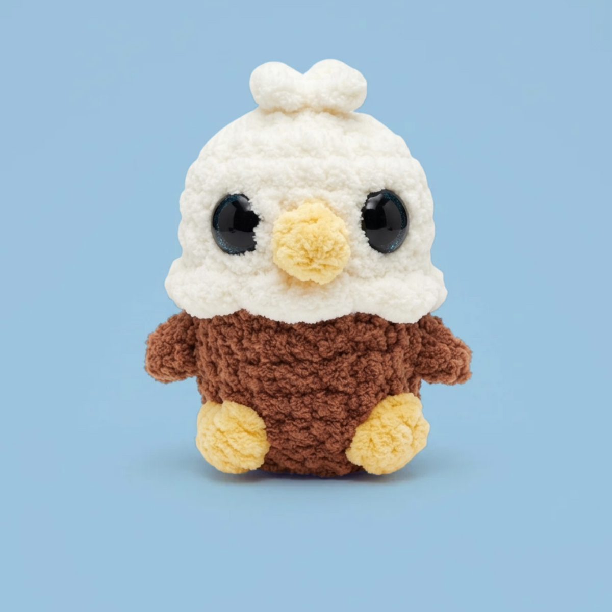 A crocheted plush toy shaped like a bald eagle, with a white fluffy head, blue eyes, a yellow beak, and a brown body with yellow feet, centered on a light blue background.