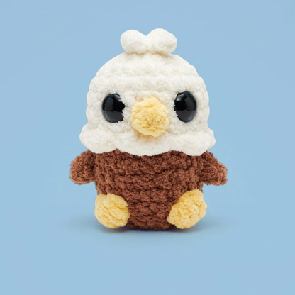 A crocheted plush toy shaped like a bald eagle, with a white fluffy head, blue eyes, a yellow beak, and a brown body with yellow feet, centered on a light blue background.