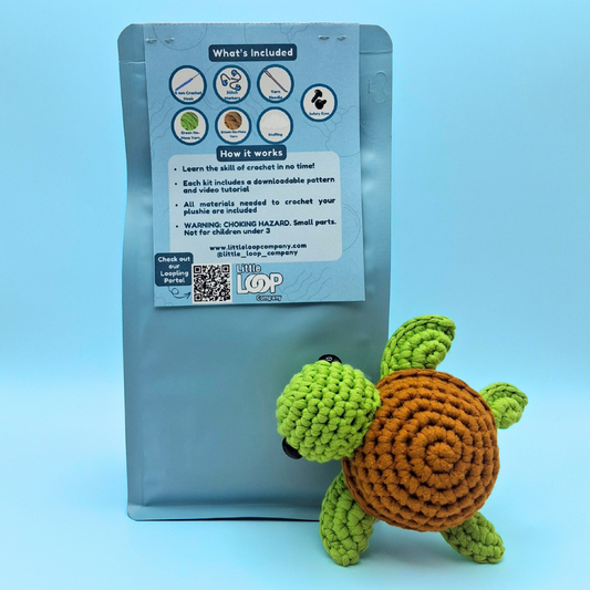 Crocheted sea turtle in front of a blue Little Loop packaging with branding and information about the kit, where to download the pattern, and what is inside the kit