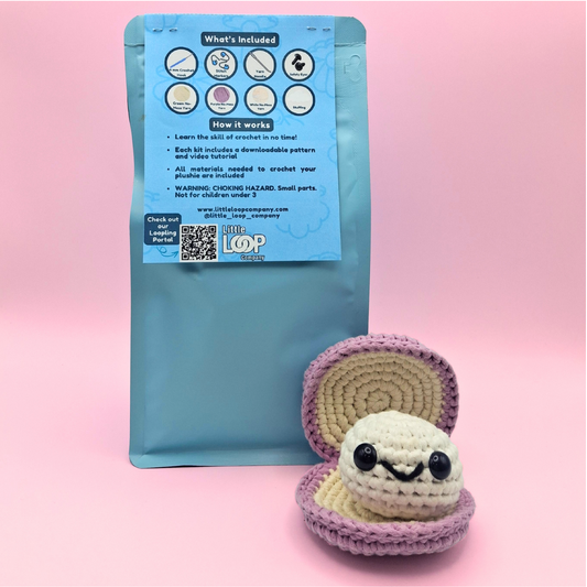 Back view of Sheldon the Clam crochet kit packaging showing included materials, step-by-step instructions, and QR code for the video tutorial, with the crocheted clam and pearl plush sitting beside the bag.