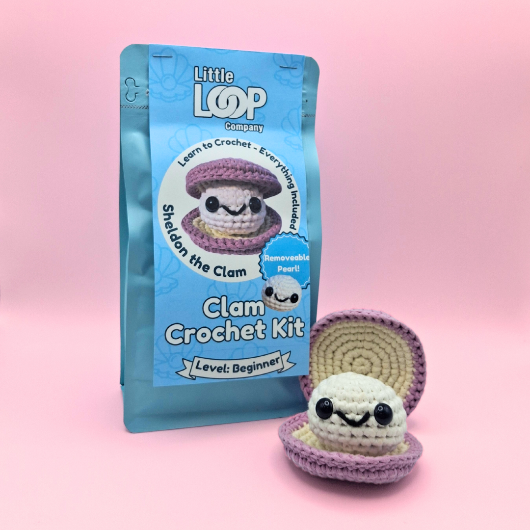 Sheldon the Clam crochet kit by Little Loop Company, featuring a cream and purple handmade clam plush with a white pearl inside, displayed in front of its resealable pastel packaging labeled ‘Clam Crochet Kit – Level: Beginner.