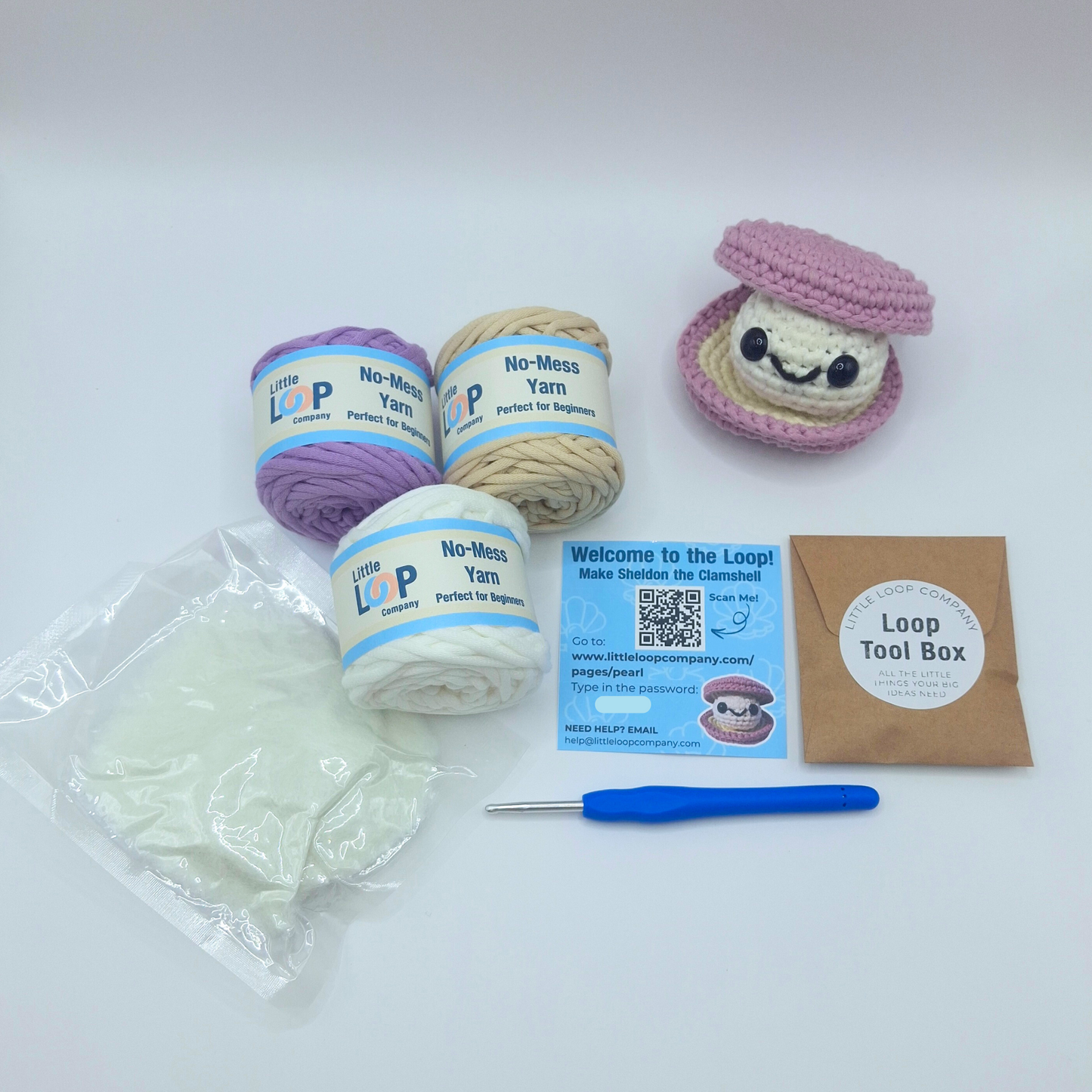Sheldon the Clam crochet kit by Little Loop Company, featuring a purple and cream crocheted clam plush with a smiling white pearl inside. Displayed with three yarn skeins in lilac, cream, and white, a crochet hook, fiberfill stuffing, a QR code instruction card, and a Loop Tool Box envelope.