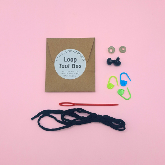 Flat lay of Little Loop Company’s Loop Tool Box supplies, including a brown kraft envelope labeled ‘Loop Tool Box,’ safety eyes, stitch markers, yarn needle, black yarn, and fiberfill stuffing arranged on a soft pink background.