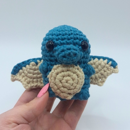 Hand holding a finished blue and beige Ember the Dragon crochet plush from Little Loop Company, showing detailed wings, snout, and stitched texture.