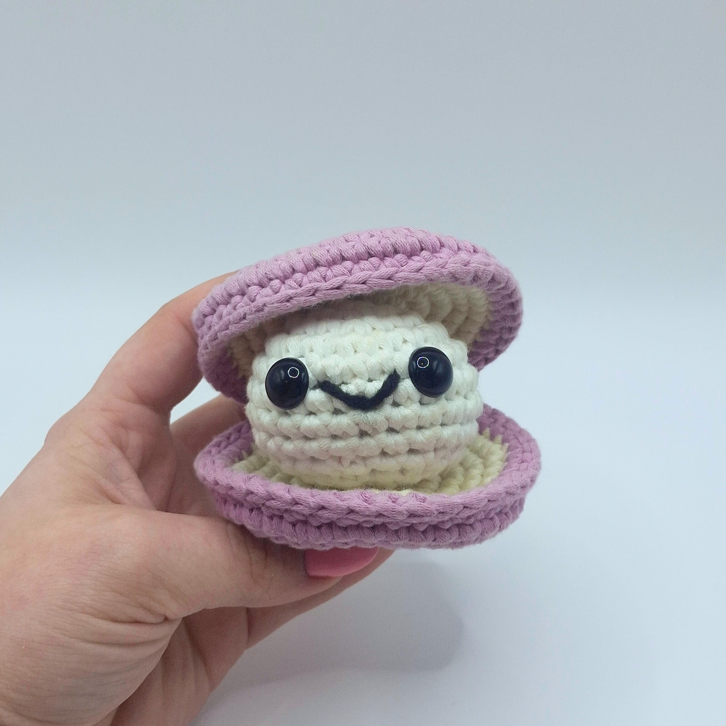 Hand holding a finished crocheted clam plush named Sheldon from Little Loop Company, with a light purple outer shell, cream inner shell, black safety eyes, and a cheerful embroidered smile.