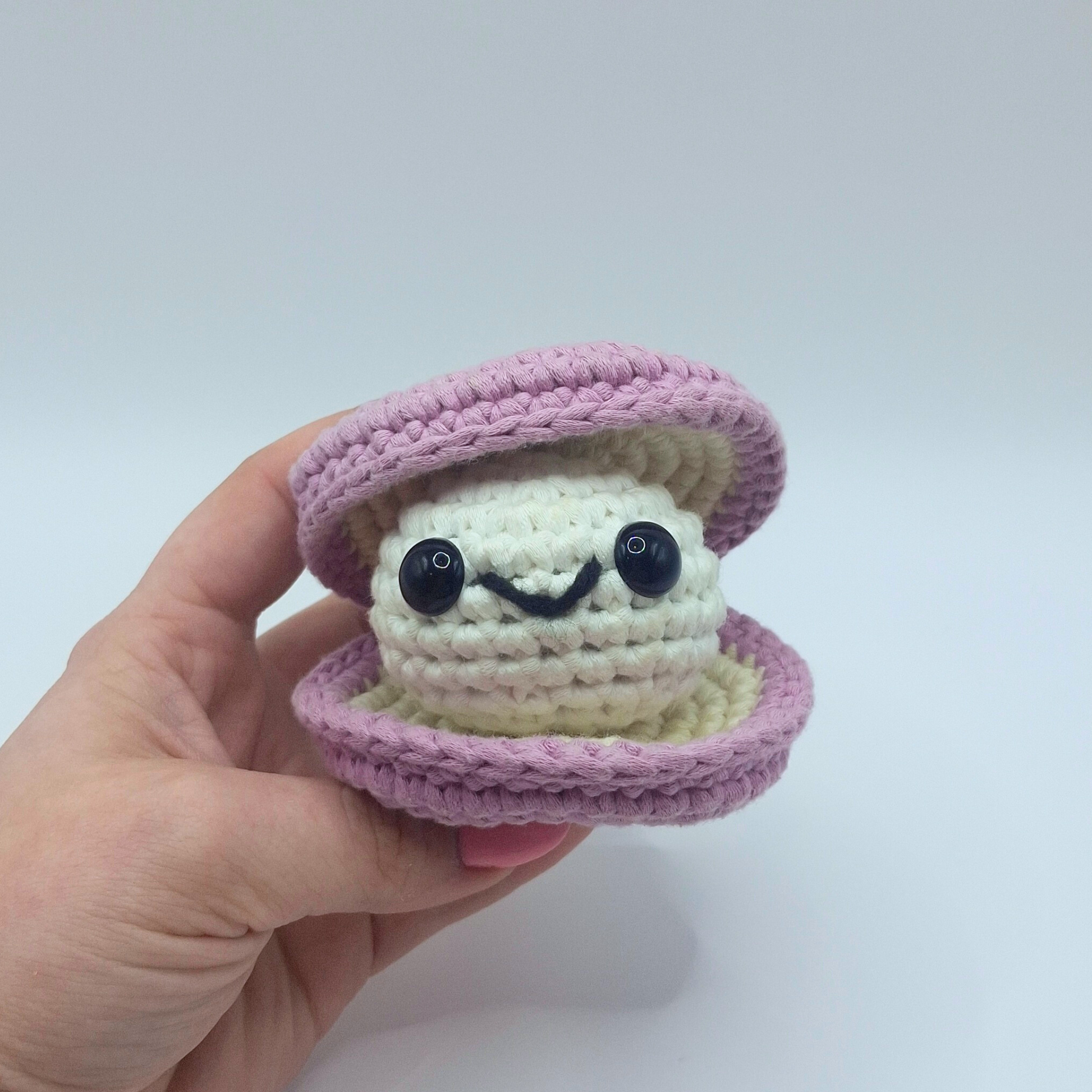 Hand holding a finished crocheted clam plush named Sheldon from Little Loop Company, with a light purple outer shell, cream inner shell, black safety eyes, and a cheerful embroidered smile.