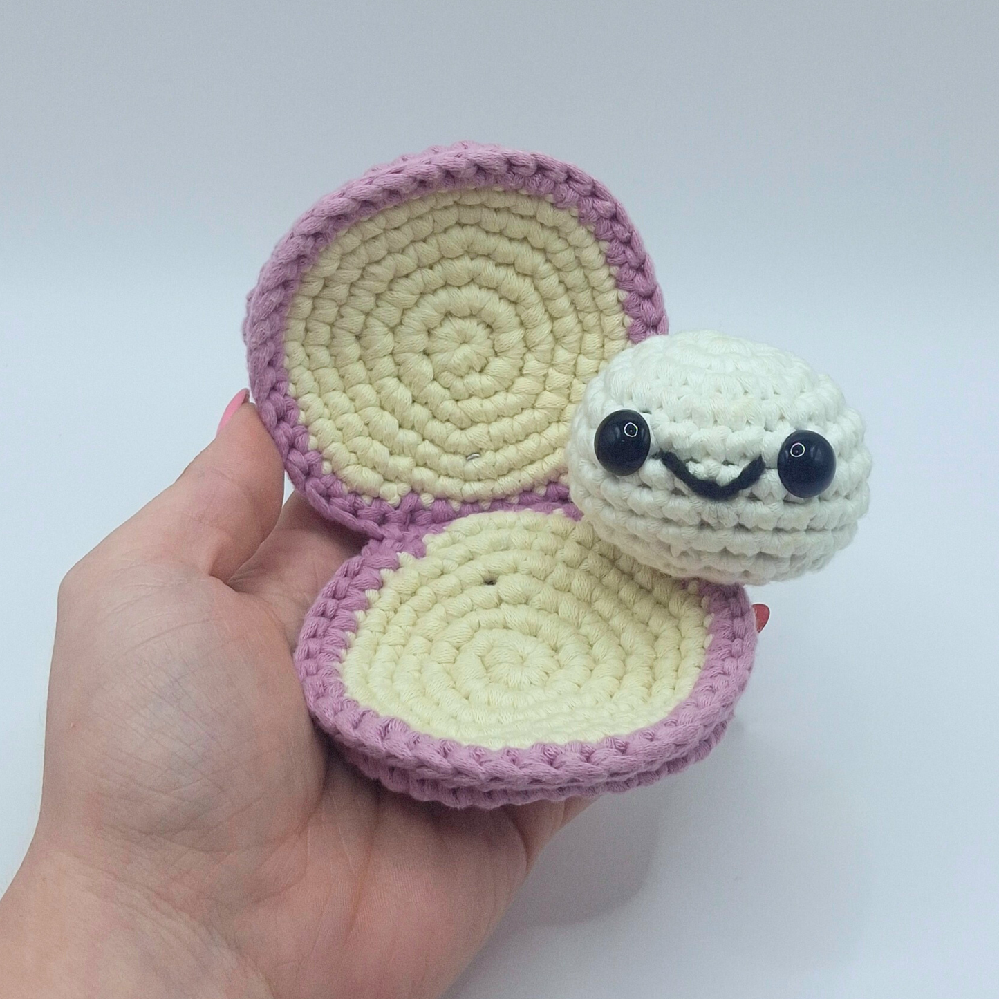 Close-up of Sheldon the Clam crochet plush with the shell open to reveal the white pearl inside. The shell is crocheted in light purple and cream yarn with a soft spiral texture, shown resting in a hand.